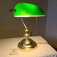 Slightly angled front view of brass library lamp on flat surface with green glass shade illuminated, shows polished brass base and curved arm with pull chain below the shade.