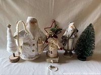 Five woodland-themed holiday decorations with two white coat Santas, one small burlap Santa, and two white cone trees with stars on wooden bases.