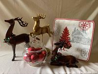 Three decorative reindeer figurines, a glass candy jar filled with matte red balls, and one holiday-themed square plate.