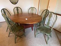 Full view of round pine table with six sage green Windsor chairs arranged around it in a carpeted room.