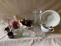 Assortment of glassware, candleholders, vases, silverplate vessels, bowl and pitcher displayed on white cloth background