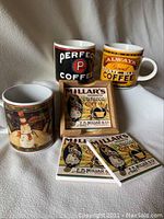 Two vintage style coffee mugs with coffee slogans and one additional mug with snowman design. Three Millar's Curacao Coffee branded coasters placed in front.