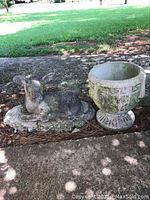 Outdoor cement deer statue lying down on an oval base beside a weathered cement planter pot, placed on a stone surface with grass background.