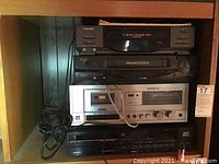 Front view of Toshiba digital VHS player, Philips Magnavox VCR Plus, Sanyo stereo cassette deck, and Magnavox CD player stacked in shelving shelf.