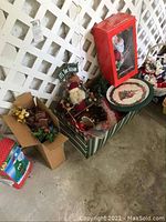 Photo showing large lot of Christmas decorations including boxed Santa Claus figurine, poinsettias, and other assorted decorations in boxes and atop a large striped box.