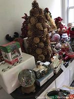 Wide view of assorted Christmas decorations on a table including pinecone tree, figurines, snow globe, and mugs