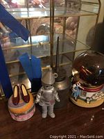 Four Wizard of Oz collectible items shown together on a shelf: metal oil can, Tin Man figure, a decorative glass case that is broken, and a music box with Dorothy and Toto next to a globe.