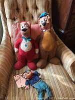 Three vintage plush toys: large Yogi Bear on right, smaller red Boo-Boo Bear on left, and Popeye marionette lying on chair seat. Chair is striped upholstered armchair.