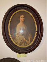 Oval framed portrait of a woman in a light off-shoulder dress with floral detail, hung on wall.