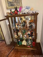 Full view of glass display cabinet containing multiple shelves filled with miniature dolls, clown figurines, animal figurines, and miniature houses.