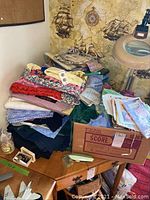 Pile of assorted fabrics, sewing papers in a box, and various sewing supplies on a wooden surface.