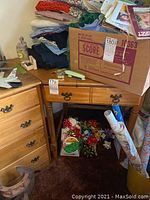 Wood corner desk surrounded by stacked fabric, gift bows, wrapping paper, and a cardboard box on top.