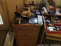 Photo of a wooden filing cabinet with drawers with multiple calculators, tape, glasses, and office items on top alongside a box of binders and shredders in the background.