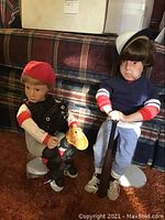 Two Danbury Mint dolls sitting on a patterned sofa; One dressed as a baseball player with a red cap and black glove, the other dressed in a navy blue jersey and sailor hat, both dolls on stands.