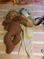 Two vintage dolls lying side by side on a striped blanket, one cloth doll with brown outfit and painted face, the other in white sleeper with pacifier.