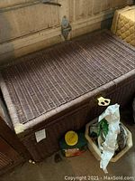 Wicker chest from front angle showing brass corners and brass hasp, small wheels visible underneath, chest placed near garage door with miscellaneous items nearby.