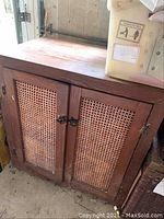 Brown wooden cabinet with two cane webbing front doors, showing top and door details, placed on a concrete floor in a garage.