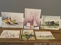Six watercolor paintings displayed on floor leaning against wall, showcasing variety in subject matter and sizes