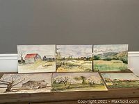 Lot of six watercolor paintings displayed on a wooden surface against a gray wall. The paintings show rural scenes including barns, trees, fields, and hills.
