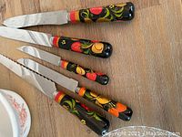 Five knives with black rounded wooden handles hand-painted in traditional Russian floral style with red, yellow, orange, green colors. Blades are a mix of straight and serrated.