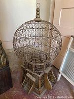 Large French metal birdcage showing detailed scrollwork with wooden base and round top finial, some dings and scratches visible.