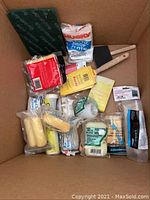 Box containing painting supplies including paintbrushes, rollers, drop cloths, sandpaper, and small hardware items.
