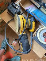 Yellow and black Dewalt electric hand sander with cord, dust collection canister attached, placed inside a cardboard box.
