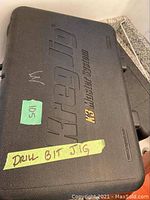 Plastic case labeled for drill bit jig with green sticker ‘105’ and yellow tape ‘DRILL BIT JIG’.