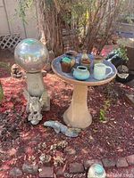 Garden setting showing the birdbath with several ceramic pots inside, orb sculpture on pedestal, gargoyle sculpture on ground, and a small lizard ceramic piece in front.