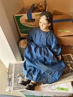 Photo of antique doll posed sitting over antique photos and two labeled cardboard boxes behind it.