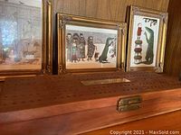 Photo showing two framed pictures with gold frames on a wood shelf, wooden box in front
