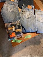 Five pairs of Levi's 501 jeans folded on wood table with plastic bag containing five pairs of new socks next to them. Jeans vary in wash from light to dark blue with branded leather patches visible on waistbands.