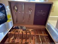 Full view of Dirilyte flatware set in original box showing various utensils including forks, knives, spoons, and serving pieces, all polished metal with mid-century modern styling.