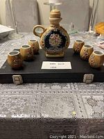 Vintage ceramic wine pitcher with stopper and six matching floral designed cups displayed on a black case over a table with patterned cloth.