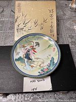 Front view of the Oriental porcelain dish displaying the hand-painted traditional Chinese female figure and scenic elements, with the original box behind.