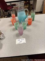 Glass beverage server with six gradient color glasses arranged around it on a table.