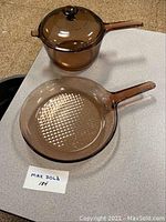 Two pieces of amber-colored Vision Corning glass cookware: a small saucepan with lid and a frying pan, both with long handles.