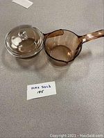Side view of transparent amber glass saucepan with handle next to matching clear lid