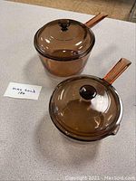 Two brown glass cookware pieces with lids, one pot and one saucepan, each with a knobbed lid and handle.