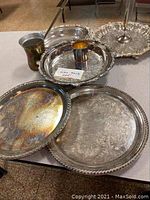 Overview of eight silver serving pieces including trays, cups, and serving dishes on beige table surface.