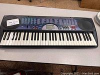 Front view of the keyboard showing full 61 piano keys, control panel with buttons, label showing 100 song bank feature, and left and right built-in speakers.