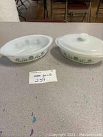 Three pieces of Glasbake glass bakeware shown on a speckled surface: two oval dishes and one lid. White glass with green leaf pattern on side dishes.