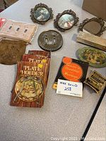 Photo showing assorted vintage items including books on plate holders, framed pictures, small pouch, booklets, and decorative items on a table.