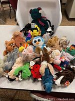 Overview of Beanie Babies plush toys spread on a white table showing various animals including bears, frogs, birds, and dogs.