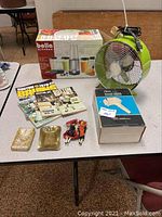 Photo showing assorted items including Bella Bliss blender box, green fan, Boston Bruins books, two small decorative trays, and two small dolls displayed on a table.