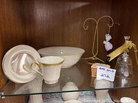 Porcelain Lenox bowl, cup, saucer with gold trim, cheese box, bell, and doll ornament arranged on shelf inside wooden cabinet.
