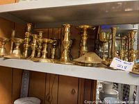 Shelf displaying multiple brass or gold-colored metal candle holders of various styles and sizes, arranged in a row on a metal shelf.
