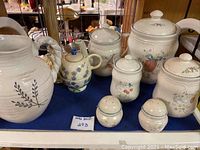 Seven ceramic kitchenware pieces with floral and fruit designs, including canisters, a pitcher, and a small tea caddy, arranged on a shelf.