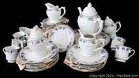 Photo of 24-piece tea set arranged with teapot, sugar bowl, creamer, 6 cups, 6 saucers, and 6 plates, showing white porcelain with blue floral pattern and gold trim