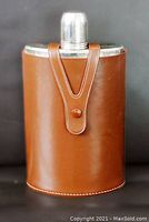 Front view of a rectangular 10oz flask covered in smooth brown leather, metal cap visible, leather strap and snap button securing the cap.